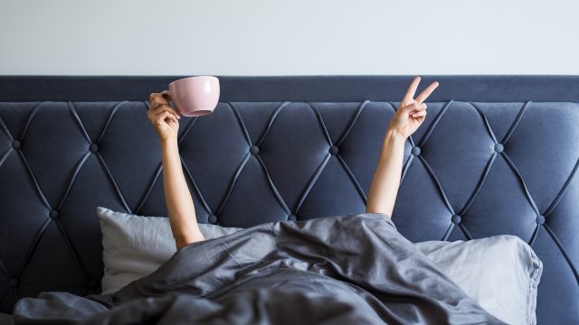 Female hands with coffee mug and victory sign sticking out from the blanket in bedroom
