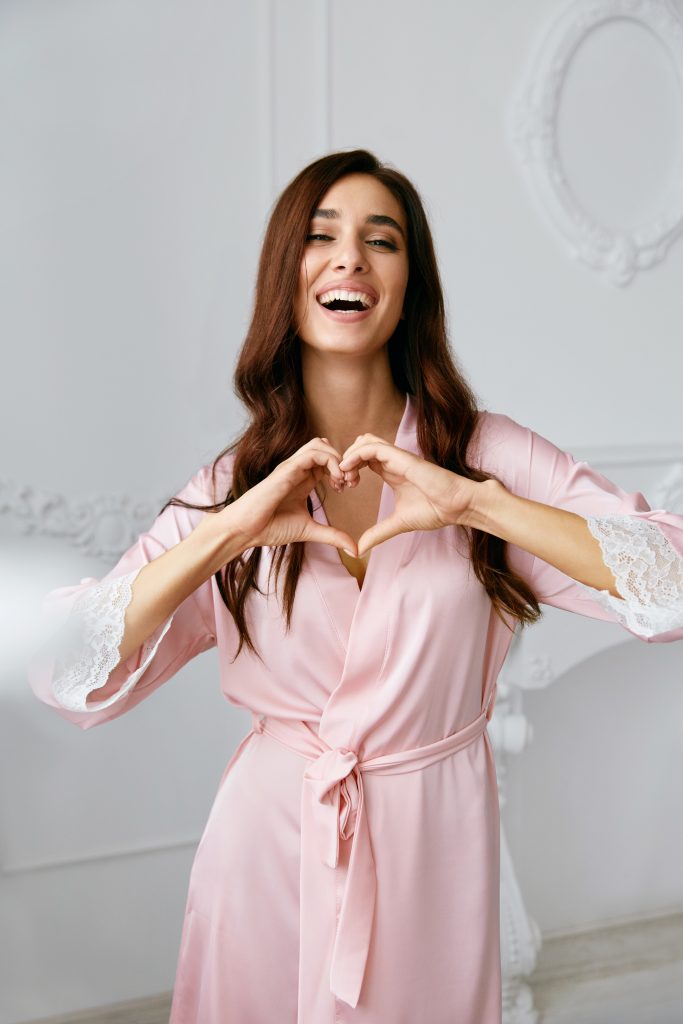 Love. Beautiful Female Model Showing Heart With Hands. Portrait Of Smiling Woman In Silk Pink Fashion Robe With Gorgeous Hair Style And Natural Makeup Making Heart Sign With Hands.