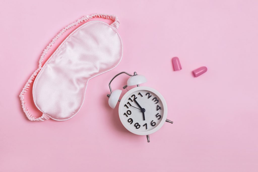 Sleeping mask, alarm clock and earplugs in pastel pink background, top view