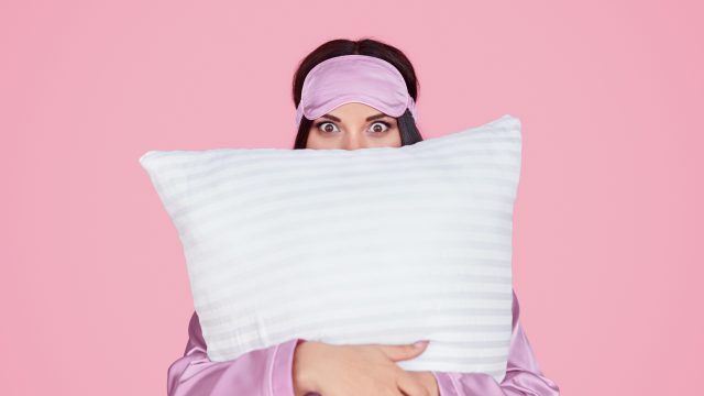 Young female in silk lilac sleep mask and pajama looking at camera and embracing comfortable pillow during bedtime against pink background. Taken just before going to sleep on silk.