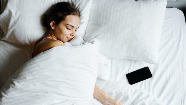Woman lying in a duvet with the sun shining in on her face and her phone next to her on the other side of the bed